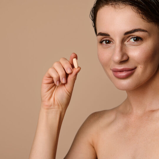 Close-up face portrait of beautiful naked woman holding a pill with dietary supplement, complex of vitamin and minerals in dragee. Beauty, skin and health care concept. Medicine, pharmacy, cosmetology