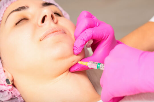 The cosmetologist makes lipolytic injection on the chin of a young woman against the double chin in a beauty salon