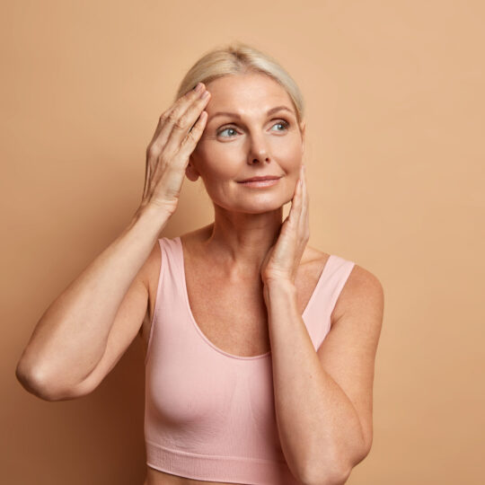 Portrait of mature elderly European woman touches face gently has perfect skin and looks thoughtfully away enjoys her soft complexion cares about appearance satisfied after anti aging procedure