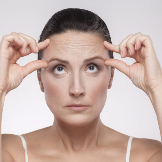 Portrait of pretty, charming, attractive middle aged woman with perfect face skin checking wrinkles on her forehead, holding fingers on brow, isolated on grey background, studio shot, copy space