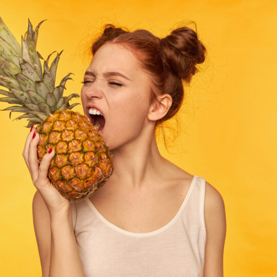 Young lady, pretty ginger woman with two buns. Wearing white shirt and trying to bite pineapple with closed eyes, healthy lifestyle. Stand isolated over yellow background